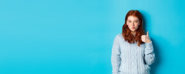 Free photo sassy redhead girl in sweater looking pleased and showing thumb up like and agree standing over blue