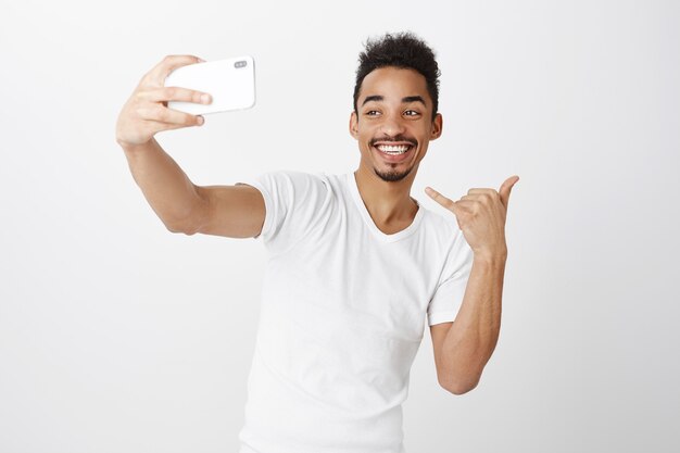 Sassy smiling african-american guy taking selfie on smartphone, showing yolo gesture