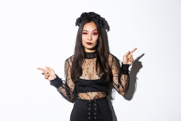 Sassy young evil witch with gothic makeup and wreath, looking arrogant while pointing fingers sideways, showing two halloween themed banners, standing over white background.