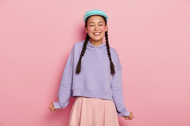 Бесплатное фото Удовлетворенная очаровательная молодая корейская модель с румяными щеками, смеется от радости, в синей кепке, большом джемпере и юбке.