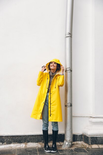 satisfied adult girl 20s wearing yellow coat standing with hood on looking up enjoying rainy weather with smile