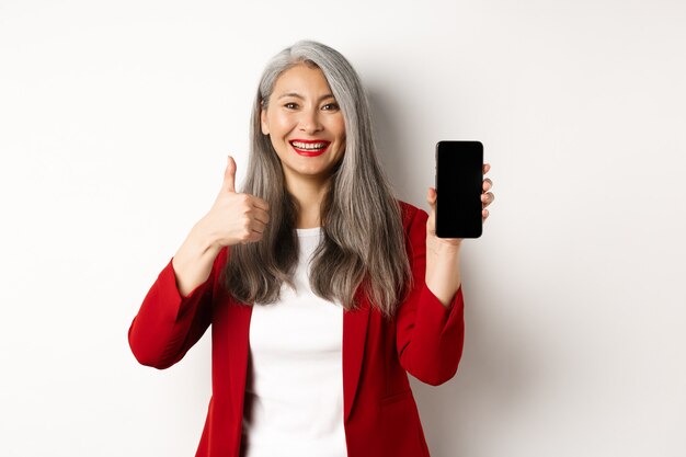 흰색 배경 위에 서서 빈 스마트폰 화면과 엄지손가락을 위로 올려 온라인 프로모션이나 회사 앱을 칭찬하는 만족스러운 아시아 노인 여성