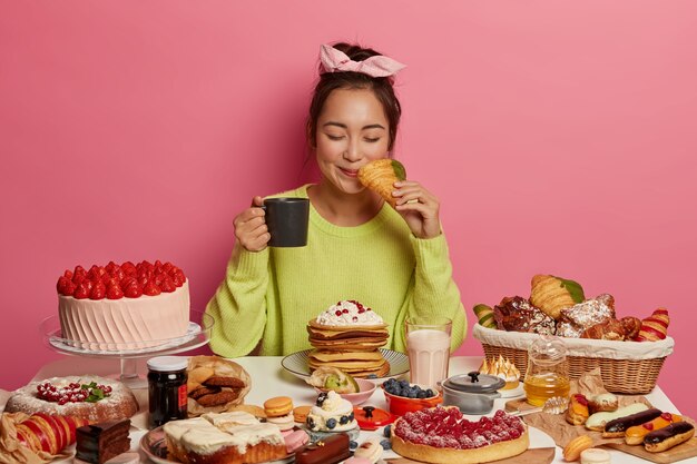 Satisfied Asian woman eats delicious croissants for every meal of day, drinks tea, poses at festive table, being addicted to sweet food, poses against pink background.