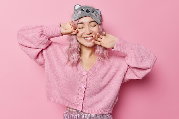 Satisfied Asian woman with dyed hair keeps eyes closed smiles gently imagines something pleasant wears sleepmask and jumper touches face gently isolated over pink background. Happy emotions concept