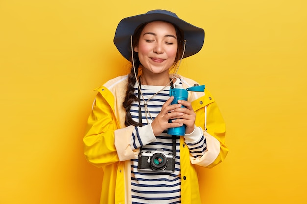 Free photo satisfied attractive female with pigtail, drinks hot coffee or tea from flask, wears casual waterproof clothes, retro camera hanging on neck, likes adventures and traveling