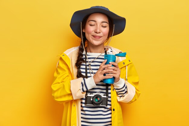 Satisfied attractive female with pigtail, drinks hot coffee or tea from flask, wears casual waterproof clothes, retro camera hanging on neck, likes adventures and traveling