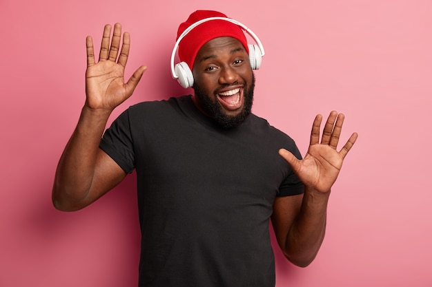 Satisfied bearded male youngster listens merry song in headphones, moves over pink background, boosts mood with cool music, feels upbeat, wears red hat and black t shirt.