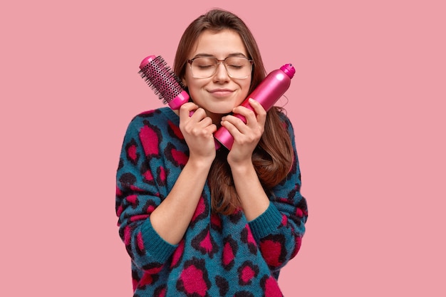 satisfied beautiful brunette lady has dreamy expression, carries hairbrush and spray for making coiffure, wears big eyewear