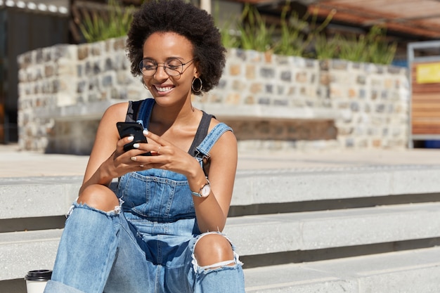 Satisfied blogger texts funny message for post on personal website, dressed in ragged overalls, sends feedback, downloads file, wears eyewear, poses at city steps, copy space for your advertising text