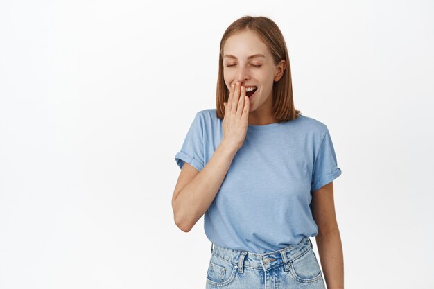 Satisfied blond woman, yawning with pleasure, had good night sleep, great nap, close eyes and smile during yawn, cover mouth with hand, standing against white wall
