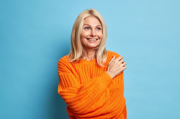 Satisfied blonde middle aged woman has dreamy expression smiles gently and thinks about something pleasant wears comfortable knitted sweater.