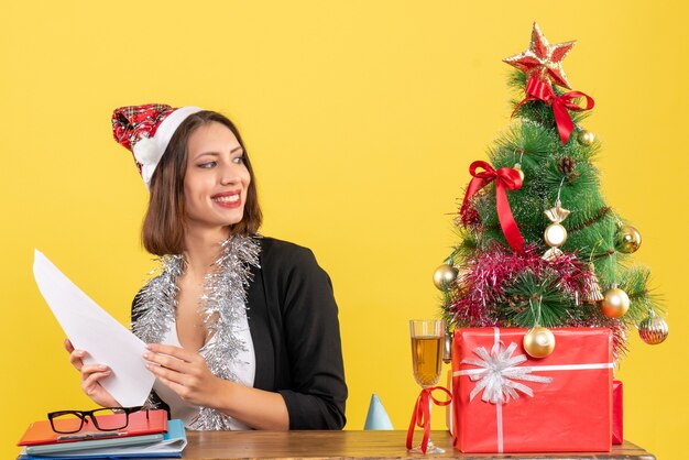 Satisfied business lady in suit with santa claus hat and new year decorations working alone and sitting at a table with a xsmas tree on it in the office