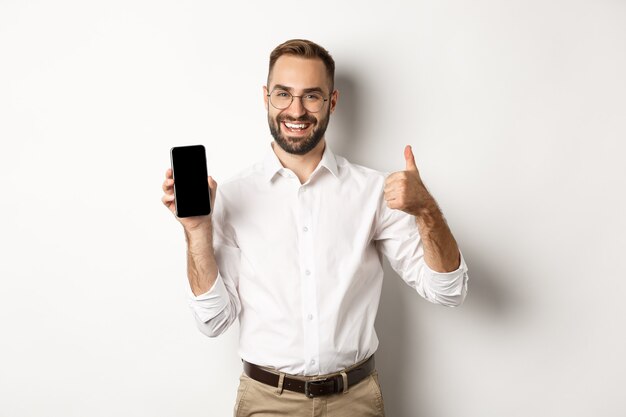 Satisfied business man in glasses showing thumbs up and demonstrating mobile phone screen, recommending app, standing over white background.