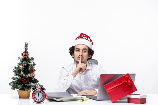 Satisfied busy proud young businessman with funny santa claus hat celebrating Christmas and making silence gesture in the office on white background