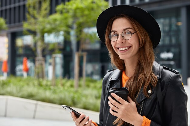 Satisfied Caucasian female checks account balance or surfes social networks, drinks coffee to go