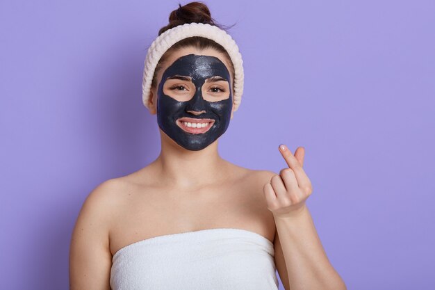 Satisfied Caucasian woman has cleansing procedures with black cosmetic mask, cleans pores, shows bare shoulders, makes korean hand sign, expresses love, makes like gesture