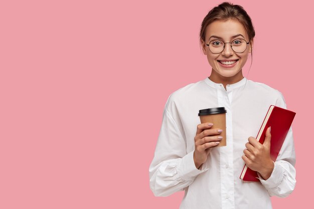 Satisfied Caucasian woman with cheerful expression, carries takeaway coffee and textbook, has positive expression