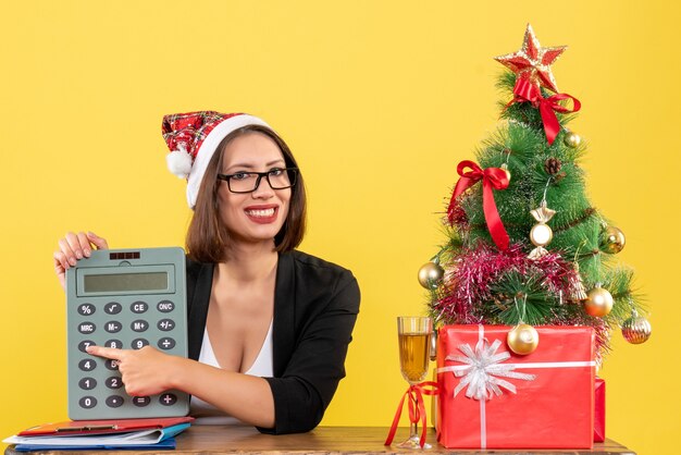 Satisfied charming lady in suit with santa claus hat and eyeglasses showing calculator in the office on yellow isolated 