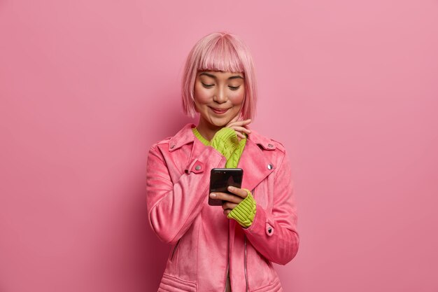 Satisfied charming woman social media blogger with stylish hairstyle, holds smartphone, reads article in internet