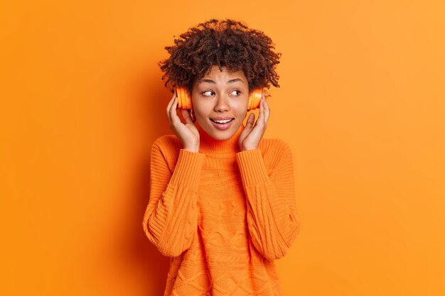 Удовлетворенная кудрявая женщина слушает аудиозапись в наушниках, смотрит в сторону, улыбается, приятно носит повседневный свитер, изолированный на оранжевой стене