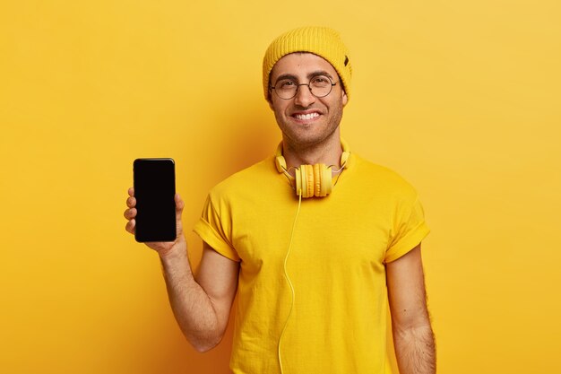 Satisfied European man holds modern cellular with black mockup space, wears yellow headgear