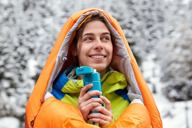 Satisfied European woman wears swimming bag, travels in snowy mountains, holds flask of hot drink