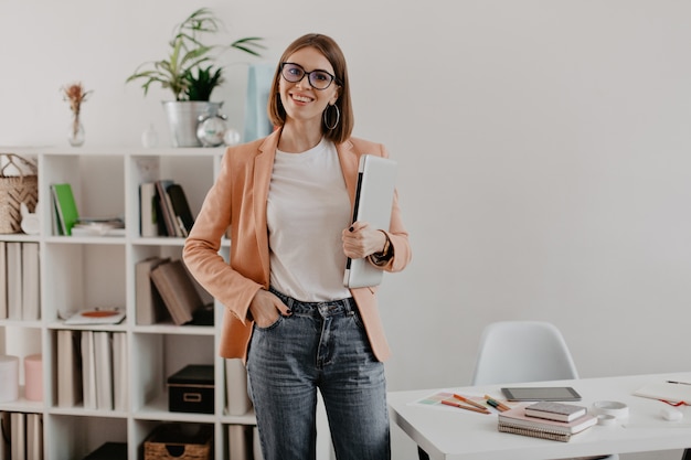 Satisfied female entrepreneur posing with laptop in hand against of her minimalistic office.