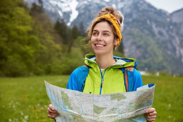 Satisfied female explorer has hitch hiking trip in mountains with snowy peaks, walks by foot on green hill, wears colorful anorak