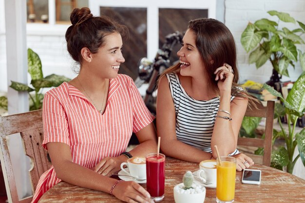 Satisfied female friends have pleasant talk together, sit at cafe, surrounded with cocktails, cappuccino and smart phone, spend leisure time in restaurant. Joyful women discuss something with fun