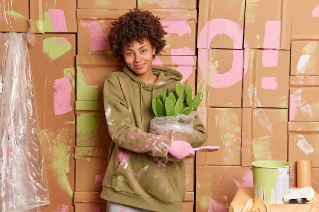 Foto gratuita la proprietaria di casa femminile soddisfatta tiene il pennello e il cactus in vaso impegnati a fare le riparazioni nella nuova casa vestita con una felpa casual che rinnova i muri con espressione felice. ristrutturazione degli interni