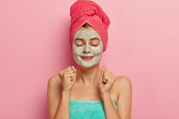Satisfied female model clenches fists with pleasure, stimulates blood circulation with help of facial mask wrapped in towel after taking shower or bath masks on regular basis has freshly cleansed face