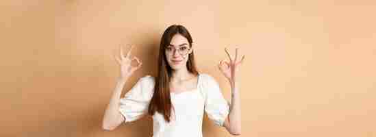 Free photo satisfied female model in glasses show okay sign looking pleased agree and approve standing on beige