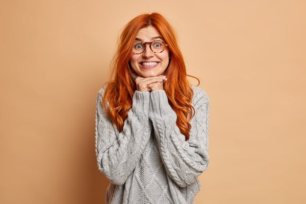 Довольная женщина-модель держит руки под подбородком взглядов с любопытной улыбкой на широко одетой в вязаный свитер.