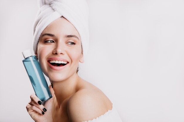 satisfied female model without makeup smiling on white wall. girl after shower posing with bottle of cosmetic tonic.