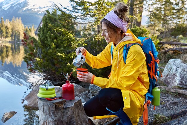 Satisfied female traveler pours coffee from coffeemaker in teacup, uses red camping butane bottle, wears raincoat with rucksack