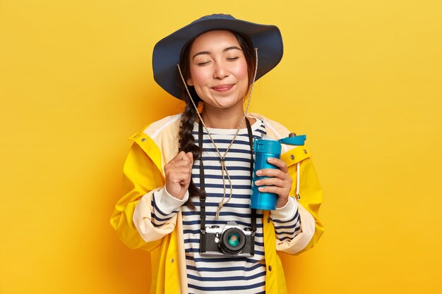 Satisfied female traveler with Asian appearance, wears hat, striped jumper and raincoat, retro camera on neck, holds flask of hot drink, isolated over yellow wall