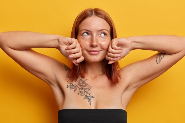 Satisfied freckled lady with natural ginger hair, touches cheeks gently, enjoys softness of skin after visiting beautician or spa salon, dressed in black dress, looks aside with dreamy expression