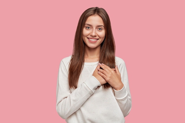 Satisfied generous brunette woman with tender smile, keeps both palms on chest