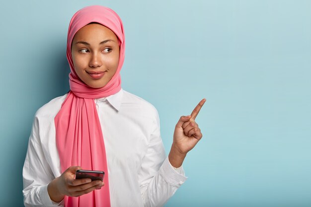 Satisfied good looking female with dark skin, holds cell phone, chats with followers in social networks, points away fore finger, demonstrates free space for promotional content, wears scarf on head