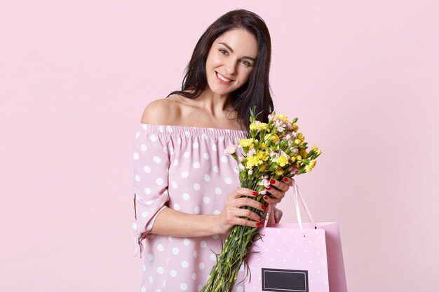 Удовлетворенная красивая женщина с темными волосами, широко улыбается, держит цветы и подарочную сумку носит летнее платье со счастливым выражением лица, модели на светло-розовом