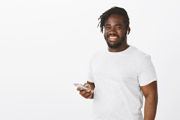 Satisfied guy with braids posing against the white wall with his phone