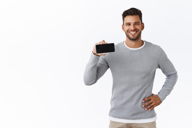 Satisfied handsome and cheerful smiling hot guy in grey sweater promote application holding smartphone horizontally stand confident casual pose advice download app try new game or visit site