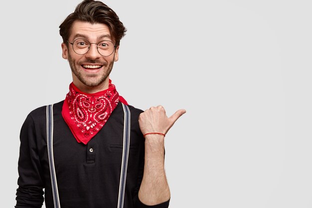 Satisfied handsome man in fashionable clothes, happy to show you direction, points aside with thumb, has joyful facial expression and charming smile