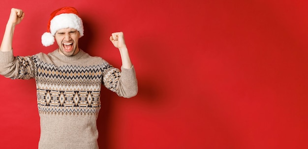 Foto gratuita soddisfatto bell'uomo che si sente fortunato e felice gridando di gioia e facendo pugni per celebrare la vittoria o la vittoria riceve un fantastico regalo di natale in piedi con il cappello di babbo natale su sfondo rosso