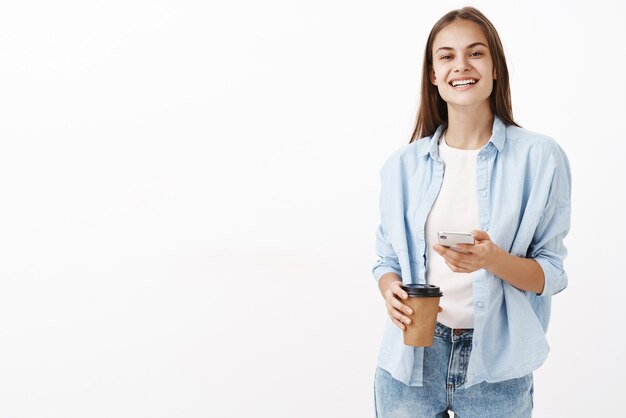 Удовлетворенная счастливая привлекательная кавказская женщина в синей блузке над футболкой держит бумажный стаканчик с кофе и мобильным телефоном, радостно улыбаясь, рада сделать перерыв во время работы в офисе