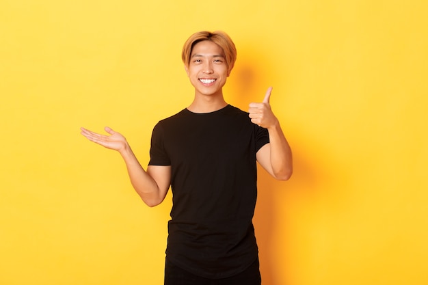 Satisfied and happy attractive korean guy smiling, showing thumbs-up in approval with rejoice, holding something on hand, standing yellow wall