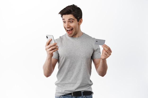 Satisfied happy man looking at smartphone screen, showing credit card, smiling excited, making online order, shopping in app, white wall
