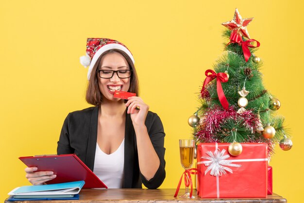 Satisfied happy smiling charming lady in suit with santa claus hat and eyeglasses showing bank card in the office on yellow isolated 