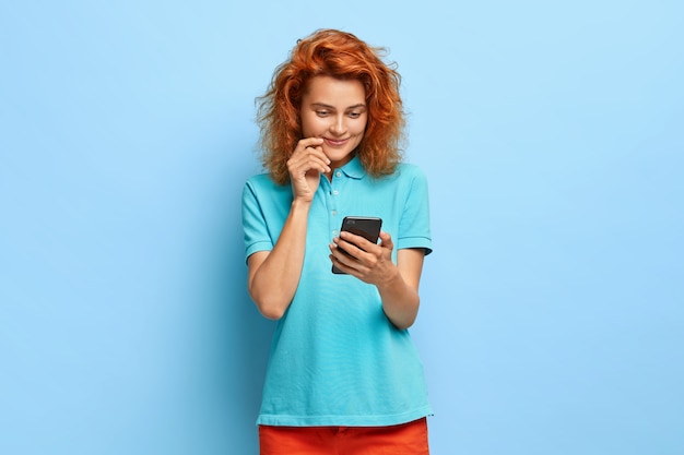 Довольная милая рыжая женщина держит мобильный телефон, рада прочитать сообщение с хорошими новостями, носит повседневную одежду, стоит у синей стены, любит пользоваться современными технологиями, у нее нежная улыбка.