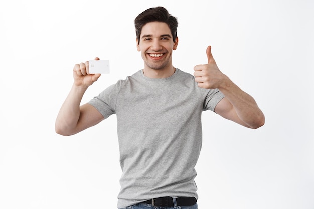 Satisfied male bank client shows thumbs up and smiles pleased, give approval, like and agree, recommend banking, standing over white background